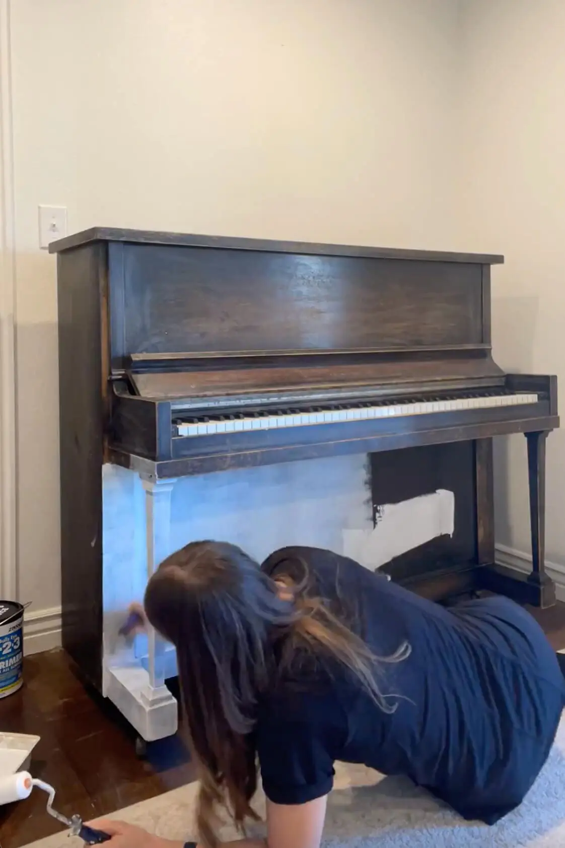 Woman applying a primer coat to a dark brown upright piano using a paint roller, preparing the surface for a fresh paint transformation.